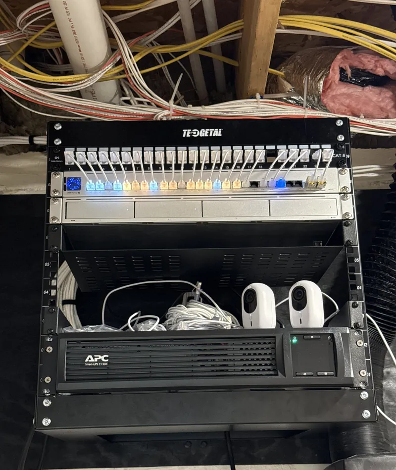 Wall rack with a patch panel, lit patch cables, a UniFi switch, a camera recorder and a battery backup